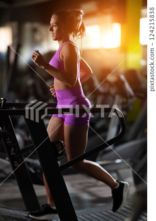 Woman Running on Treadmill in Gym.A fit woman wearing a purple sports outfit is running on a treadmill in a gym. She has a focused yet relaxed expression, with gym equipment and a warm light in the Woman Running on Treadmill in Gym.A fit woman wearing a purple sports outfit is running on a treadmill in a gym. She has a focused yet relaxed expression, with gym equipment and a warm light in the 124215838