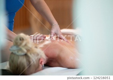 Relaxing Back Massage in a Spa Setting. A professional massage therapist in a blue uniform provides a relaxing back massage to a client lying on a massage table. 124216051