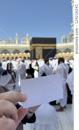 A pilgrim holds a small notebook with a heart doodle in front of the Kaaba in Mecca, Saudi Arabia. The sacred site is surrounded by worshippers performing Hajj or Umrah under a clear blue sky 124216241