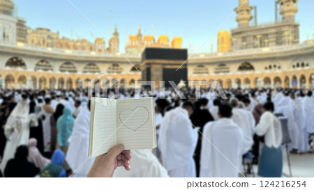 A pilgrim holds a small notebook with a heart doodle in front of the Kaaba in Mecca, Saudi Arabia. The sacred site is surrounded by worshippers performing Hajj or Umrah under a clear blue sky 124216254