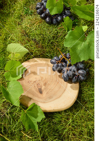 Top view of bunches of grapes with dew drops on their shiny skins placed on a fresh lawn. Vine with leaves, covering one side of a circular wooden tray in the center. Space to place products 124216674