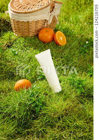 An unlabeled tube of cosmetics sits tilted on a green lawn with lush foliage. A ripe orange sits around it. The orange is cut in half and placed next to a bamboo picnic basket with a lace border. 124216709