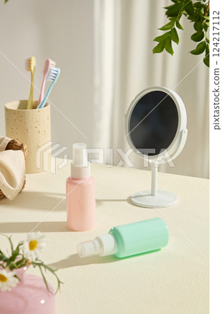 Two unlabeled spray bottles displayed on white table. A round plastic standing mirror. Colorful toothbrushes placed in a cup. Decorated by green branches. Health and natural care cosmetics concept. Two unlabeled spray bottles displayed on white table. A round plastic standing mirror. Colorful toothbrushes placed in a cup. Decorated by green branches. Health and natural care cosmetics concept. 124217112