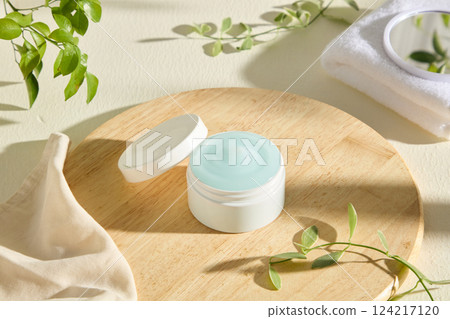 An open cosmetic jar filled with light blue cream is placed in the center, on a light beige round wooden tray. The jar lid is tilted next to it. Beige cheesecloth and leaves decorated around. An open cosmetic jar filled with light blue cream is placed in the center, on a light beige round wooden tray. The jar lid is tilted next to it. Beige cheesecloth and leaves decorated around. 124217120