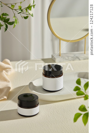 Two plastic jars placed on a marble tray which displayed on a white surface. A round mirror in the right corner of the photo, green leaves decorate the surrounding sides. Bottle mockup for cosmetics 124217128