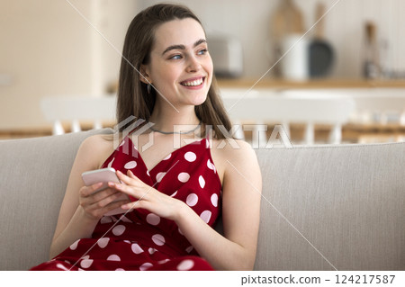 Serene young girl holding cellphone on couch looking at distance Serene young girl holding cellphone on couch looking at distance 124217587