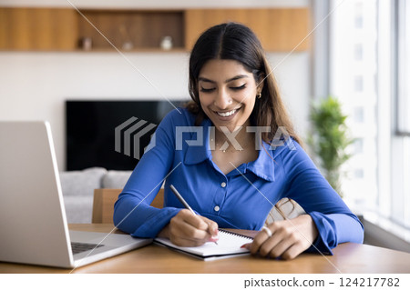 Woman smiles, writes in notebook seated at desk with laptop Woman smiles, writes in notebook seated at desk with laptop 124217782