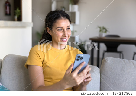 Cheerful woman using smartphone seated on couch at home 124217862