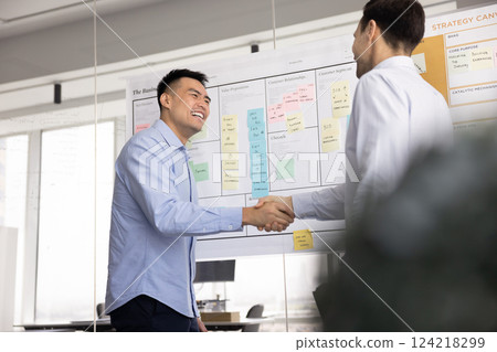 Cheerful diverse male business partners shaking hands at whiteboard Cheerful diverse male business partners shaking hands at whiteboard 124218299