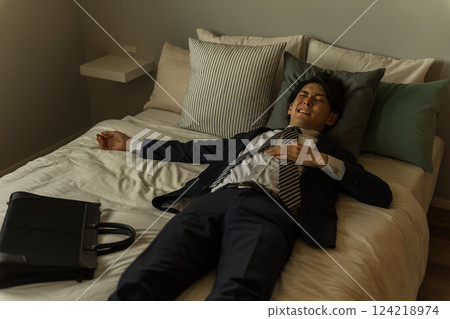 一位年輕的亞洲男商人回家後穿著西裝躺在床上(加班、過勞、公司員工、健康狀況不佳) 一位年輕的亞洲男商人回家後穿著西裝躺在床上(加班、過勞、公司員工、健康狀況不佳) 124218974