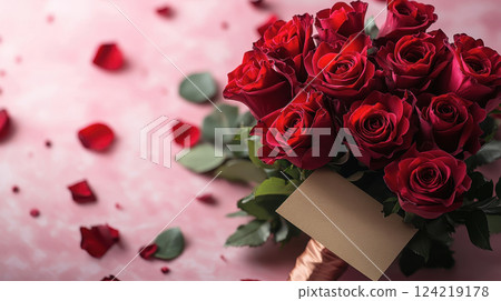 A stunning bouquet of velvety red roses with lush green leaves, wrapped elegantly and paired with a blank card. Scattered rose petals on a soft pink background complete the romantic aesthetic. 124219178