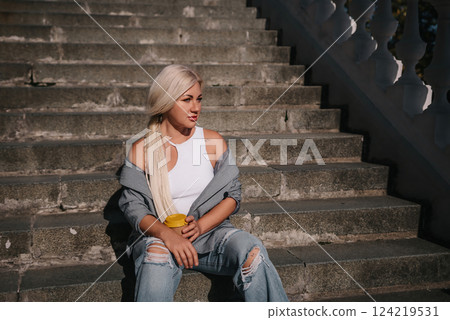 A blonde woman sits on a set of stairs, holding a cup. She is wearing a gray jacket and blue jeans. 124219531