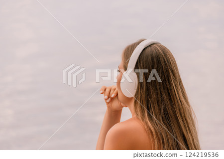 A woman wearing headphones is looking out at the water. The scene is calm and peaceful, with the woman seemingly lost in thought. 124219536