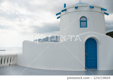 A white building with blue doors and windows. The building is located near the ocean. The sky is cloudy and the building is surrounded by mountains. A white building with blue doors and windows. The building is located near the ocean. The sky is cloudy and the building is surrounded by mountains. 124219604