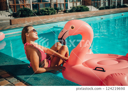 A woman in a pink bikini is sitting on a pink inflatable flamingo in a pool. The scene is playful and fun, with the woman enjoying her time in the water. A woman in a pink bikini is sitting on a pink inflatable flamingo in a pool. The scene is playful and fun, with the woman enjoying her time in the water. 124219698