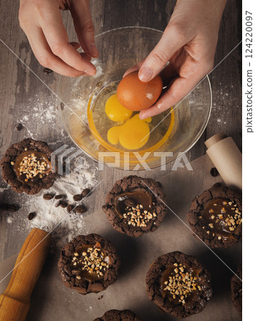 Female hands break eggs into a bowl next to muffins on baking paper on a table, top view, vertical image. Female hands break eggs into a bowl next to muffins on baking paper on a table, top view, vertical image. 124220097