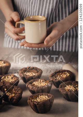 Female hands hold coffee mug over freshly baked muffins on baking paper on a table, vertical image. Female hands hold coffee mug over freshly baked muffins on baking paper on a table, vertical image. 124220099