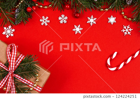 Christmas decorative border with green spruce, holly berries, snowflakes, candy cane and craft giftbox on red background, top view, copy space. 124224251