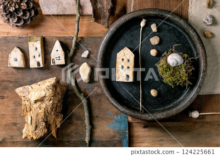 Flat lay creative design rustic composition with miniature ceramic houses, pebbles, shell, moss, pine cone, feather, twigs on wooden background. Natural materials arrangement. Flat lay creative design rustic composition with miniature ceramic houses, pebbles, shell, moss, pine cone, feather, twigs on wooden background. Natural materials arrangement. 124225661