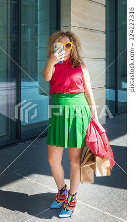 a beautiful young woman in bright clothes with a phone and bags takes selfies or takes pictures of the city on a walk 124227316