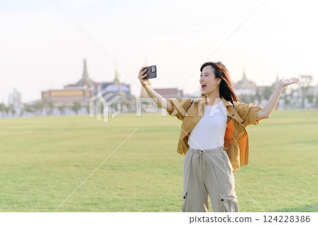 Traveler asian woman in her 30s making a livestream or selfie on smartphone while explores Wat Phra Kaew emerald Buddha. Share the wonders of Thai heritage through her journey. Traveler asian woman in her 30s making a livestream or selfie on smartphone while explores Wat Phra Kaew emerald Buddha. Share the wonders of Thai heritage through her journey. 124228386