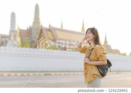 Traveler asian woman in her 30s, backpack slung over her shoulder, explores the intricate details of Wat Pra Kaew with childlike wonder. Sunlight dances on the golden rooftops. 124228401