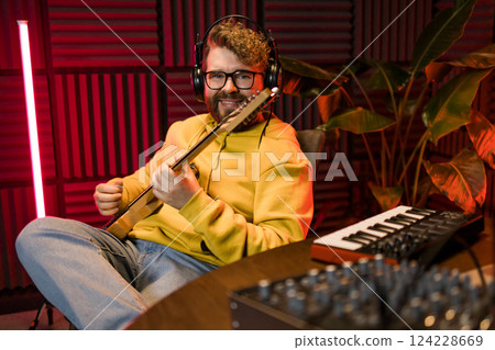 A focused male musician fine-tunes a guitar against the backdrop of a digital sound editing setup in a contemporary recording studio environment. 124228669