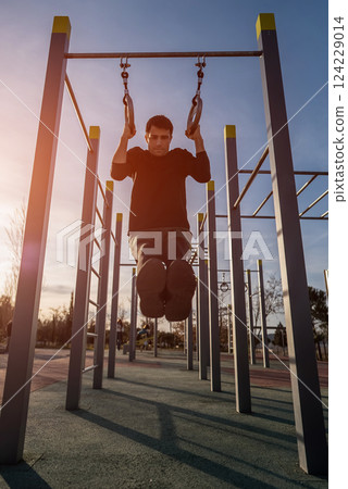 young athletic man exercising rings at fitness calisthenics workout and making pull-ups at street gym during sunset, body improvement and strength training concept 124229014