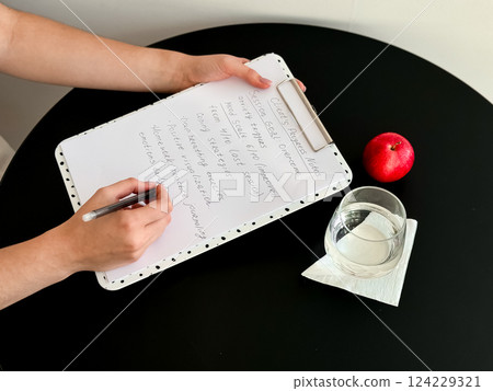 Female writing on clipboard with notes next to apple and glass on round black table 124229321