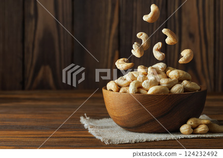 Cashew nuts falling into bowl placed on small rustic jute tablecloth, creating a visually appealing arrangement against dark wooden background 124232492