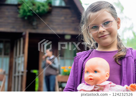 Smiling Young Girl in Glasses Holding a Doll Outdoors in Front of a Wooden House in the Countryside 124232526