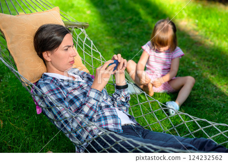 Mother relaxing in a hammock using a smartphone while her young daughter sits on the grass playing with her shoes in a sunny backyard, depicting family leisure and digital distraction outdoors Mother relaxing in a hammock using a smartphone while her young daughter sits on the grass playing with her shoes in a sunny backyard, depicting family leisure and digital distraction outdoors 124232562