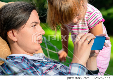 Mother relaxing in a hammock using a smartphone while her curious daughter leans in to see the screen, depicting family interaction and digital technology in an outdoor setting. Mother relaxing in a hammock using a smartphone while her curious daughter leans in to see the screen, depicting family interaction and digital technology in an outdoor setting. 124232563