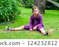Young Girl in a Purple Outfit Sitting on Grass in a Green Garden, Looking Thoughtful and Curious 124232812