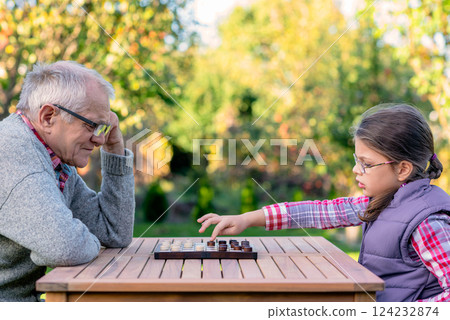 Elderly grandfather and young granddaughter playing checkers together at a wooden table in a garden, enjoying quality family time and strategic thinking outdoors in autumn Elderly grandfather and young granddaughter playing checkers together at a wooden table in a garden, enjoying quality family time and strategic thinking outdoors in autumn 124232874