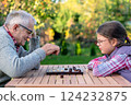 Elderly grandfather and young granddaughter playing checkers together at a wooden table in a garden, enjoying quality family time and strategic thinking outdoors in autumn 124232875