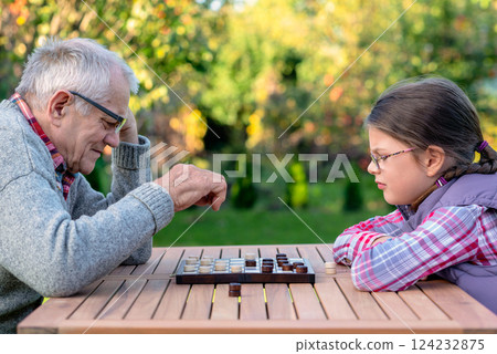 Elderly grandfather and young granddaughter playing checkers together at a wooden table in a garden, enjoying quality family time and strategic thinking outdoors in autumn 124232875