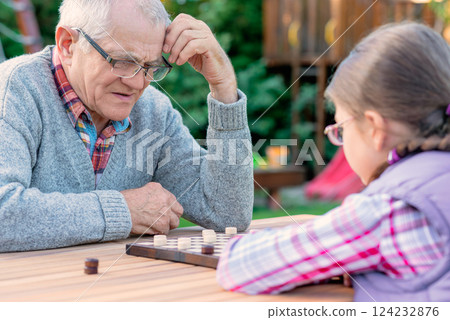 Elderly grandfather and young granddaughter playing checkers together at a wooden table in a garden, enjoying quality family time and strategic thinking outdoors in autumn 124232876