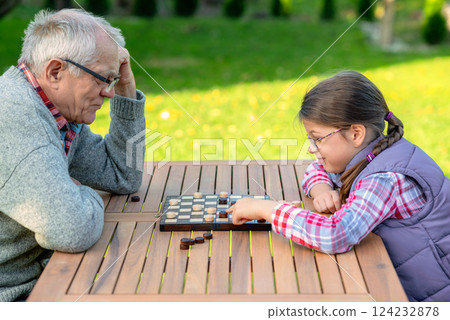 Elderly grandfather and young granddaughter playing checkers together at a wooden table in a garden, enjoying quality family time and strategic thinking outdoors in autumn 124232878