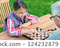 Young girl playing checkers with her grandfather at a wooden table outdoors, focusing on strategy and enjoying quality family time in a park or garden setting 124232879