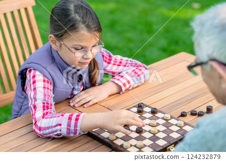 Young girl playing checkers with her grandfather at a wooden table outdoors, focusing on strategy and enjoying quality family time in a park or garden setting Young girl playing checkers with her grandfather at a wooden table outdoors, focusing on strategy and enjoying quality family time in a park or garden setting 124232879