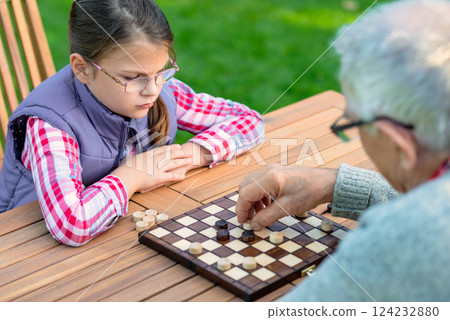 Young girl playing checkers with her grandfather at a wooden table outdoors, focusing on strategy and enjoying quality family time in a park or garden setting 124232880