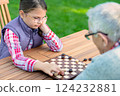 Young girl playing checkers with her grandfather at a wooden table outdoors, focusing on strategy and enjoying quality family time in a park or garden setting 124232881