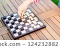 Close-up of a hand moving a piece on a wooden checkers board during an outdoor game, focusing on strategy and decision-making 124232882