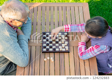 Elderly grandfather and young granddaughter playing checkers together at a wooden table in a garden, enjoying quality family time and strategic thinking outdoors in autumn Elderly grandfather and young granddaughter playing checkers together at a wooden table in a garden, enjoying quality family time and strategic thinking outdoors in autumn 124232883