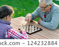 Elderly grandfather and young granddaughter playing checkers together at a wooden table in a garden, enjoying quality family time and strategic thinking outdoors in autumn 124232884