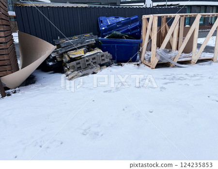 Construction debris stacked beside a black metal fence on a snowy urban street. Includes blue dumpsters, concrete slabs, wooden frames, and discarded materials Construction debris stacked beside a black metal fence on a snowy urban street. Includes blue dumpsters, concrete slabs, wooden frames, and discarded materials 124235853