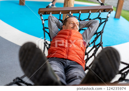 Man in bright orange vest relaxing on hammock outdoors. Lying back with hands behind head, appearing content, with tranquil expression. Comfortable outdoor leisure Man in bright orange vest relaxing on hammock outdoors. Lying back with hands behind head, appearing content, with tranquil expression. Comfortable outdoor leisure 124235974