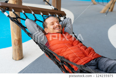 Relaxed man smiling contentedly while resting in hammock, wearing vibrant orange vest. His hands are behind his head, showcasing state of peacefulness and joy 124235975