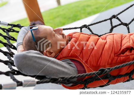 Man in orange vest comfortably resting in hammock outdoors. Appears relaxed and calm, hands behind head, sunglasses on forehead. Capturing a moment of leisure and tranquility 124235977